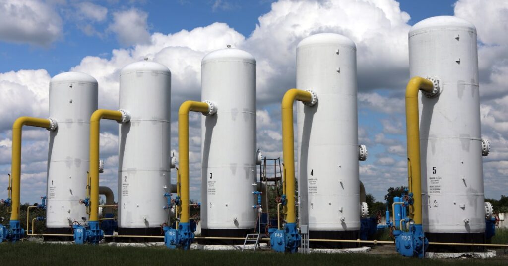 Numerous gas boosters, tanks, and pipes are neatly lined up in a vast field beneath a partly cloudy sky.