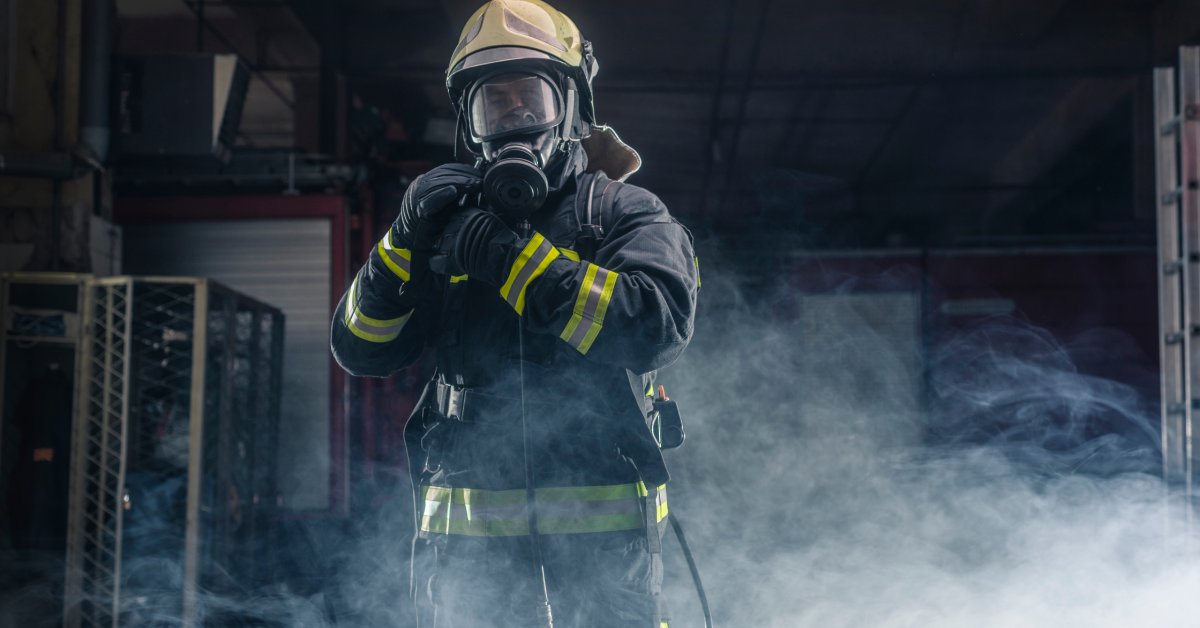 A firefighter in full protective gear, including a helmet and breathing apparatus, moves through a smoke-filled building.