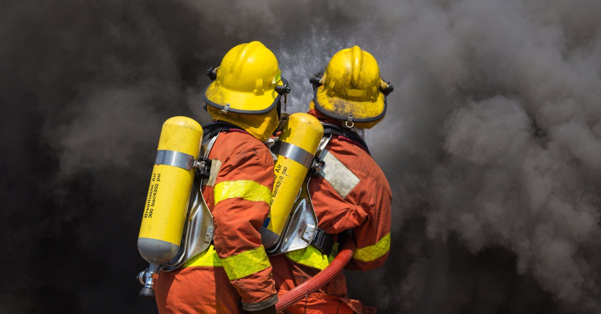 Two firefighters wearing heavy oxygen tanks on their backs hurry into a dense cloud of thick, dark smoke.